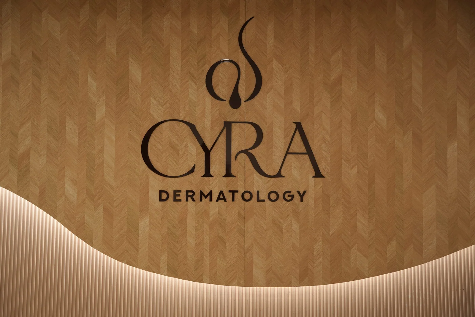 The Cyra Dermatology logo on a herringbone timber wall above a softly lit curve